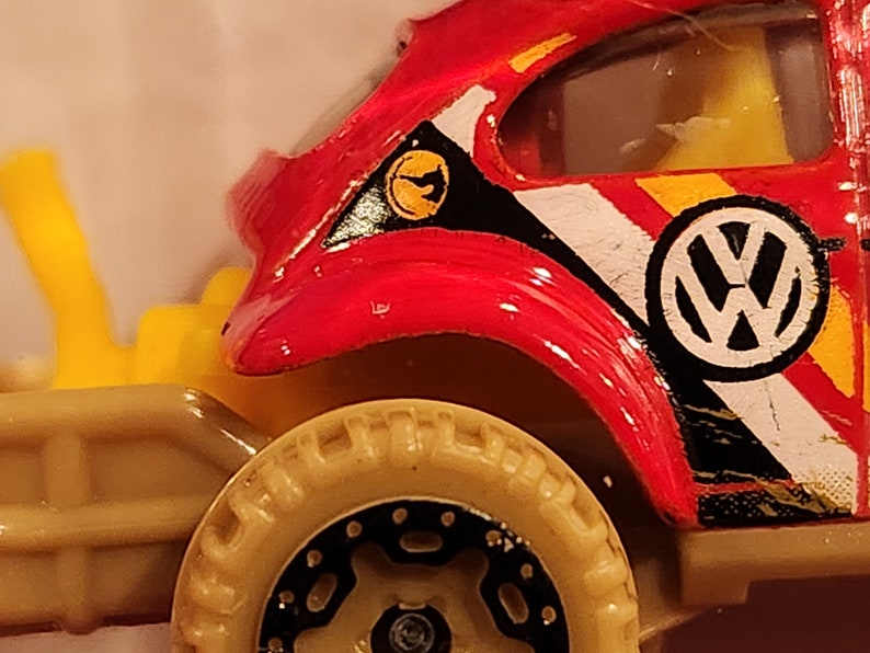May include: Red, black and white toy car with a Volkswagen logo. The car has a yellow and black stripe on the side and a yellow and black decal on the hood. The car has a black and tan tyre.