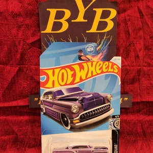 Peut inclure: Une voiture miniature Hot Wheels de couleur violette avec des finitions blanches et un toit blanc. La voiture est sur une carte bleue et blanche avec le logo Hot Wheels et les mots "Rod Squad" imprimés dessus. La voiture est numéro 320/250.