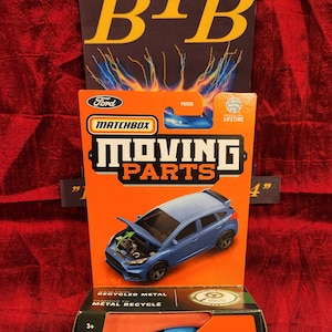 Könnte beinhalten: Ein Matchbox Moving Parts Modell eines blauen Ford Focus RS, mit geöffneter Motorhaube. Die Verpackung ist orange und schwarz, mit dem Ford-Logo und dem Text "MOVING PARTS". Eine zweite Box zeigt das Auto in zwei Blautönen.