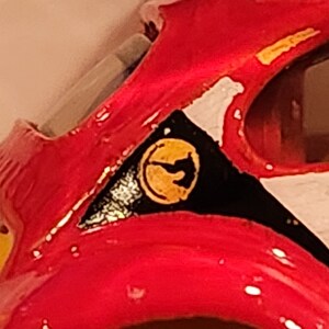 May include: Close-up of a red toy car with a black and white stripe design. The car has a yellow logo with a stylised bird design.