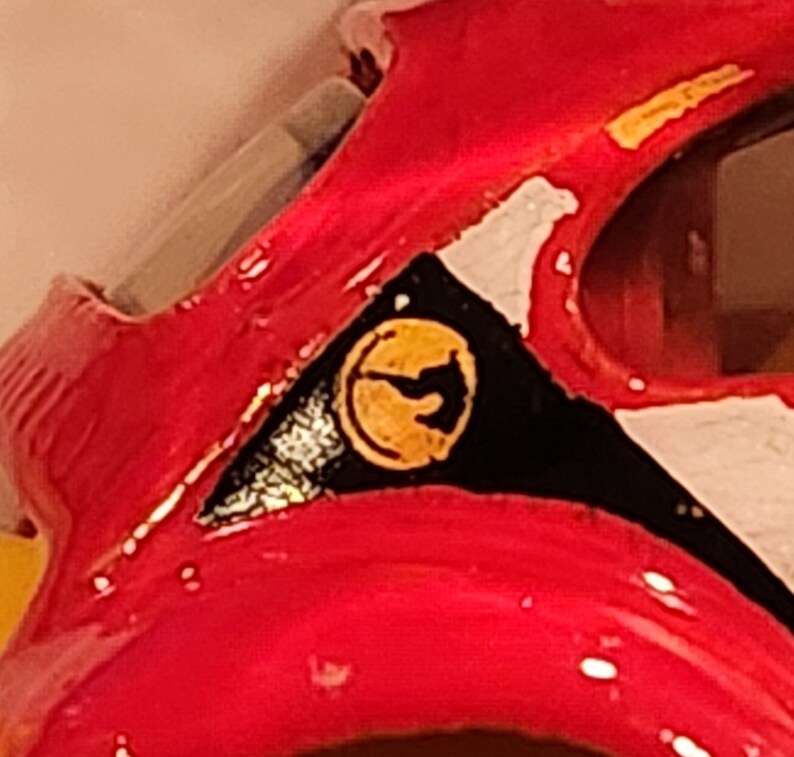 May include: Close-up of a red toy car with a black and white stripe design. The car has a yellow logo with a stylised bird design.