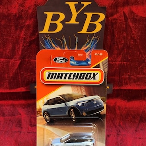 Könnte beinhalten: Ein blaues Matchbox-Spielzeugauto aus Metall, das einen Ford Mustang Mach-E darstellt. Das Auto befindet sich auf einem weißen Hintergrund mit einer Stadtstraßenkulisse im Hintergrund. Die Verpackung trägt das Matchbox-Logo und die Worte "Ford" und "07/125".