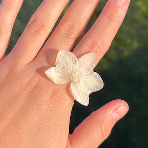 Op de afbeelding: Een ring met een groot, doorschijnend, gebroken wit bloemmotief met delicate nerven op de bloemblaadjes en een klein, rond accent in het midden. De bloem is gemaakt van een lichtgewicht materiaal en valt op als sieraad aan de vinger.