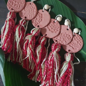 Puede incluir: Una colección de adornos redondos de color terracota con patrones en relieve similares al encaje, cada uno emparejado con una borla de hilo rojo, blanco y verde. Los adornos están ensartados con un cordón blanco y cuentas de madera, sobre una gran hoja verde.