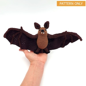 Puede incluir: Un murciélago de crochet marrón con alas grandes, una nariz pequeña y una sonrisa burlona. El murciélago está sostenido en la mano de una persona.
