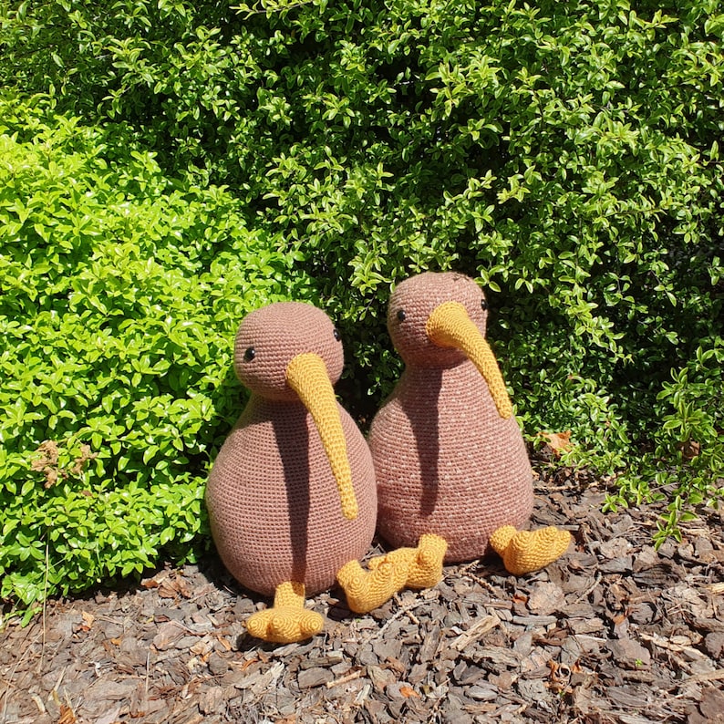 May include: Two brown knitted kiwi bird plushies with yellow beaks and feet. The plushies are sitting on a bed of brown mulch.