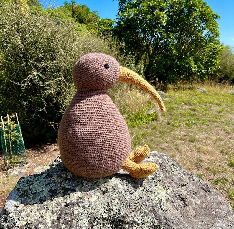 May include: A brown crocheted kiwi bird with a yellow beak and feet sitting on a rock. The kiwi is a popular symbol of New Zealand.