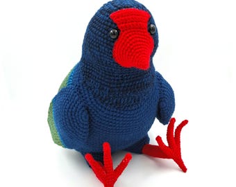 Peut inclure: Un oiseau crocheté à la main, dans des tons de bleu, de rouge et de vert. L'oiseau a un bec et des pieds rouges, avec des yeux noirs. Le texte "PATTERN ONLY" est dans un rectangle jaune en haut à droite. L'oiseau est assis.