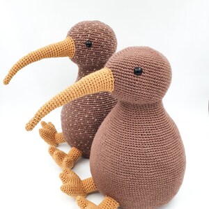 May include: Two brown crocheted kiwi bird plushies with orange beaks and feet. The kiwi on the right is larger than the one on the left.