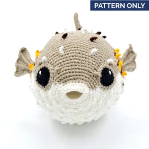Peut inclure: Un jouet de poisson-globe crocheté dans les tons beige, blanc et noir. La peluche faite à la main présente de grands yeux noirs, de petites épines marron et des accents jaunes sur les nageoires. Le texte "PATTERN ONLY" est dans un rectangle bleu.