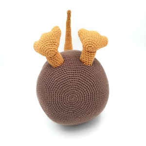 May include: A brown crocheted kiwi bird toy with orange feet and a long tail. The toy is sitting on a white surface.