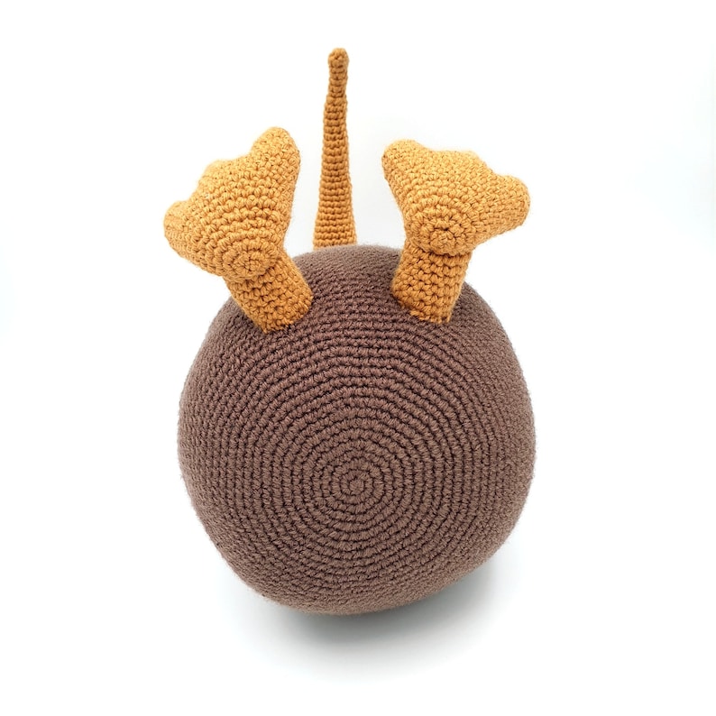 May include: A brown crocheted kiwi bird toy with orange feet and a long tail. The toy is sitting on a white surface.