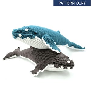 May include: Two crocheted whale plushies, one blue and one grey, with white accents. The whales are stacked on top of each other, with the blue whale on top. The whales have realistic features, including eyes, mouths, and blowholes.