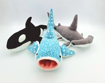 May include: Three crocheted stuffed animal sharks: a black and white orca, a blue whale shark with white spots, and a gray hammerhead shark.