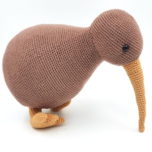 May include: A brown crocheted kiwi bird toy with a long orange beak and black eyes. The toy is standing on a white surface.