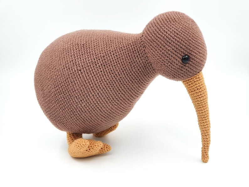 May include: A brown crocheted kiwi bird toy with a long orange beak and black eyes. The toy is standing on a white surface.