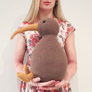 May include: A brown crocheted kiwi bird plush toy with a yellow beak and feet. The toy is being held by a person wearing a floral dress.
