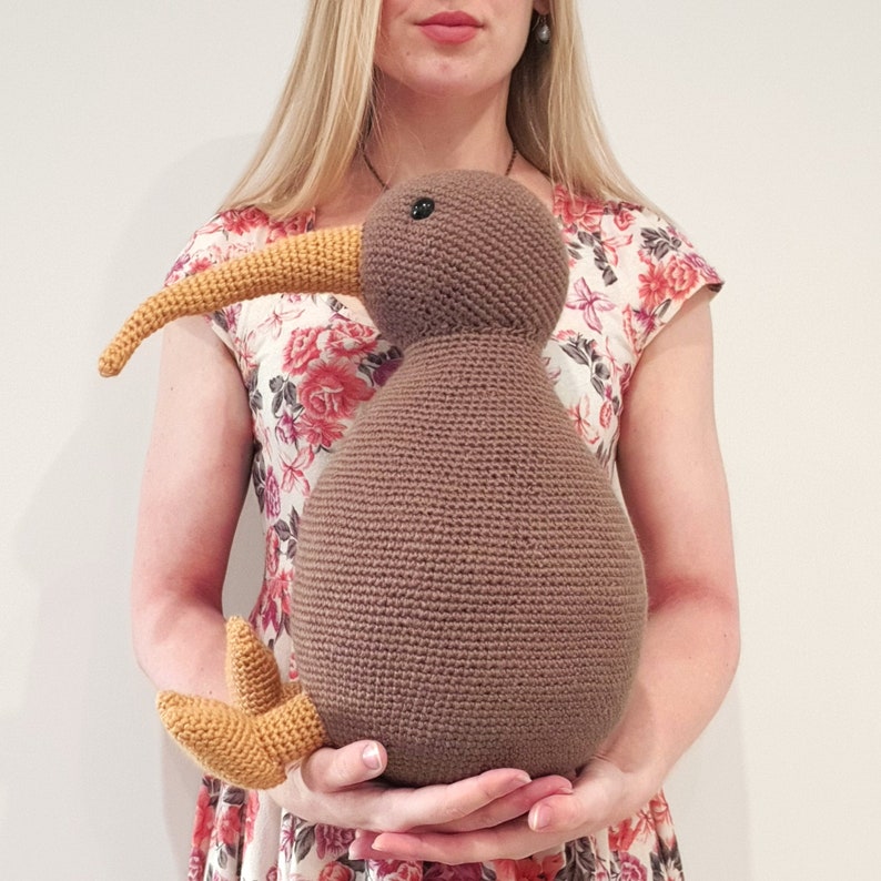 May include: A brown crocheted kiwi bird plush toy with a yellow beak and feet. The toy is being held by a person wearing a floral dress.