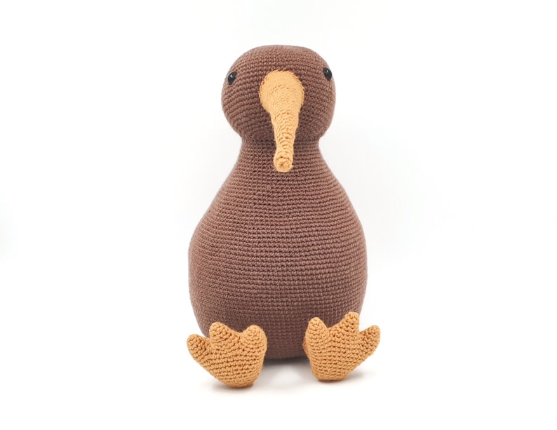 May include: A brown crocheted kiwi bird plush toy with orange feet. The toy has black eyes and a long orange beak.