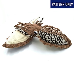 May include: Crocheted cuttlefish in shades of brown, cream, and white. The cuttlefish has a detailed pattern on its body and a large, reflective eye. The text "PATTERN ONLY" is in a blue rectangle in the upper right corner.