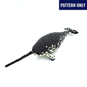 May include: A crocheted narwhal toy in grey and white, with a long, spiralled tusk. The body is dark grey with white spots, and the tail and flippers are also white. The text "PATTERN ONLY" is in a blue rectangle.