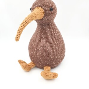 May include: A brown and tan crocheted kiwi bird plush toy with black eyes. The kiwi is sitting with its legs outstretched.