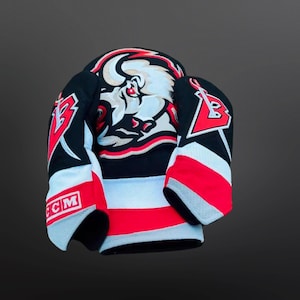 Puede incluir: Una camiseta de hockey negra y roja con un logotipo de búfalo blanco en la parte delantera. La camiseta tiene rayas blancas y rojas en las mangas y un logotipo de CCM en la manga izquierda.