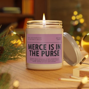 Puede incluir: Una vela de cera de soja encendida en un tarro de cristal con tapa plateada. La etiqueta dice "MERCE IS IN THE PURSE" y "REAL HOUSEWIVES OF BEVERLY HILLS". La vela está perfumada con especias, azúcar moreno, canela, limón, vainilla y pralinés.