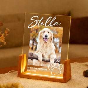 Könnte beinhalten: Ein beleuchteter Gedenk-Fotorahmen mit dem Namen "Stella" in Schreibschrift. Der Rahmen zeigt ein Foto eines Golden Retrievers, Pfotenabdrücke und den Text "IN LOVING MEMORY 2001-2023". Der Rahmen besteht aus Holz und hat einen warmen Schein.