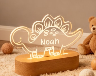 Könnte beinhalten: Ein Dinosaurier-Nachtlicht mit dem Namen "Noah" in Weiß beleuchtet. Das Licht steht auf einem Holzsockel und ist von dekorativen Kugeln und einem Teddybären umgeben. Entworfen für ein Kinderzimmer.
