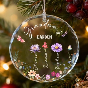 Może przedstawiać: Przezroczysta szklana bombka choinkowa z napisem "Grandma's Garden" elegancką czcionką. Ozdoba zawiera kwiatowe i motylkowe wzory, z imionami "Olivia", "Luna" i "Sophia" kursywą. Wisi na białym sznurku.
