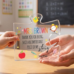 May include: A clear acrylic puzzle piece plaque featuring the text MRS. BROWN THANK YOU FOR BEING SUCH AN IMPORTANT PIECE OF MY story and the name Sophia. The design includes colourful illustrations of a book, alarm clock, apple, pen, and leaf.
