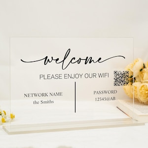 Può includere: Un cartello trasparente in acrilico con il testo "welcome" in un font corsivo. Il cartello include anche il testo "Please enjoy our wifi" e un codice QR. Sotto il testo, c'è una sezione per il nome della rete e la password. Il nome della rete è "the Smiths" e la password è "12345@AB".