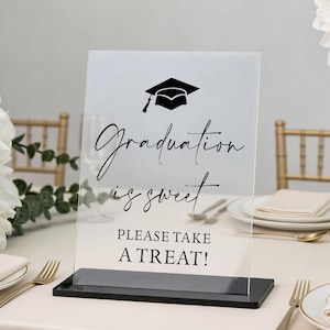 May include: Clear acrylic graduation sign with a black base. The sign features a graduation cap graphic and the words "Graduation is sweet, Please take a treat!" in elegant script. The sign is set on a table with gold silverware and white plates.