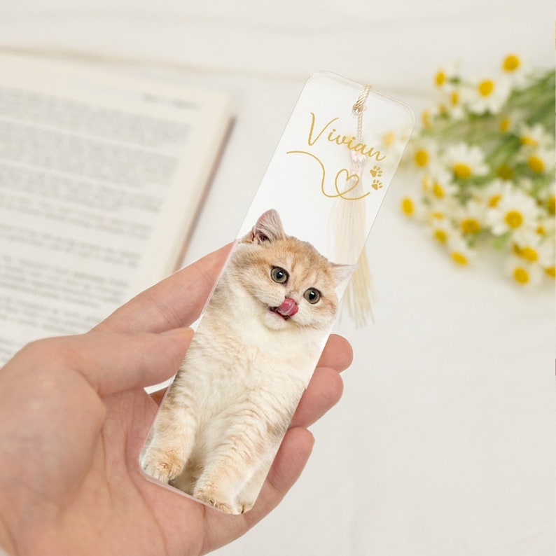 May include: A clear acrylic bookmark with a photo of a cat with its tongue sticking out. The bookmark has a gold tassel and the name "Vivian" written in gold script.