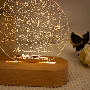 May include: A personalized night light with a constellation design. The clear acrylic disc is engraved with names "Maria & Fabian", date, and coordinates. The base is made of wood and emits a warm glow.