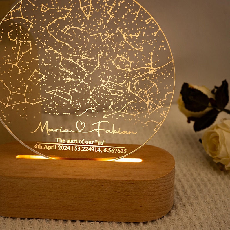 May include: A personalized night light with a constellation design. The clear acrylic disc is engraved with names "Maria & Fabian", date, and coordinates. The base is made of wood and emits a warm glow.