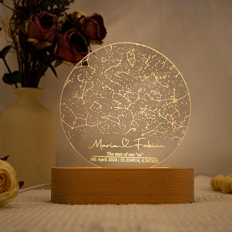 May include: A personalized night light featuring a constellation map design. The clear, circular acrylic panel is illuminated, displaying stars and connected lines. The base is made of light-colored wood. Text includes names, a date, and coordinates.