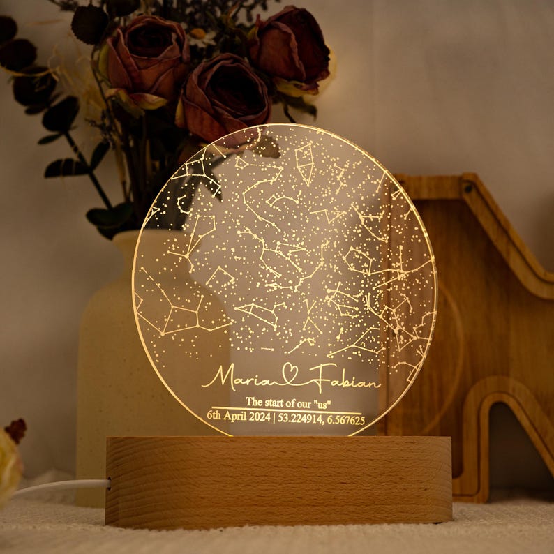 May include: A personalized night light featuring a clear, circular acrylic panel with a constellation design and the names "Maria & Fabian." The base is made of light-colored wood. The text reads "The start of our "us" 6th April 2024 | 53.224914, 6.567625."