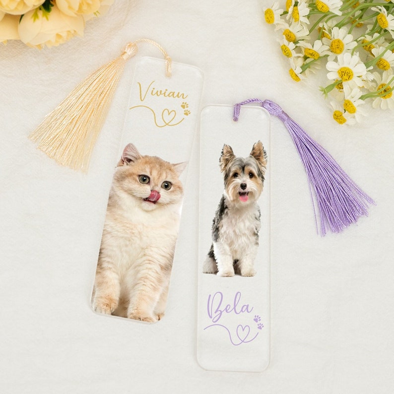 May include: Two personalized glass bookmarks with a cat and a dog on them. The cat bookmark has a cream tassel and the text "Vivian" with a heart and paw prints. The dog bookmark has a purple tassel and the text "Bella" with a heart and paw prints.