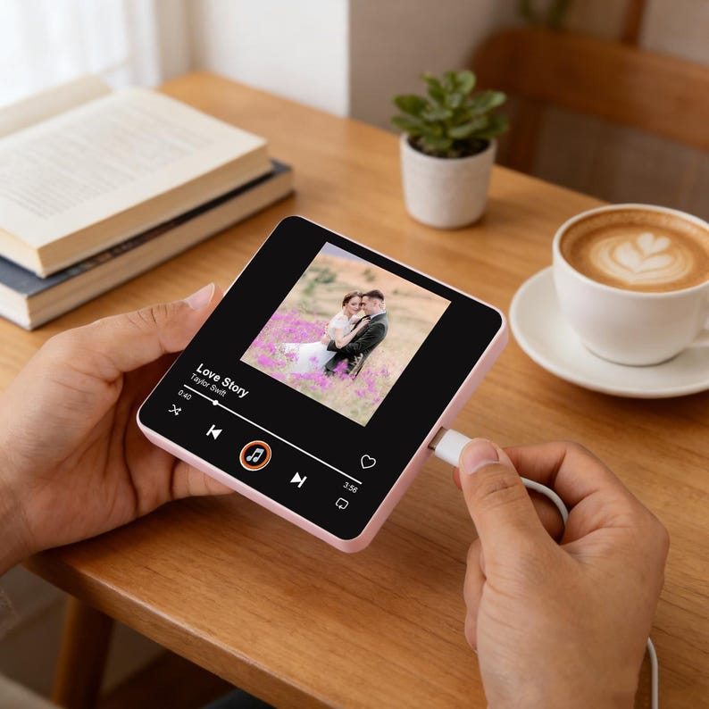 May include: A pink digital music player showing a wedding photo and the song "Love Story" by Taylor Swift. The player is connected to a white charging cable. A coffee cup, a small plant, and books are in the background.