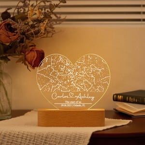 May include: A heart-shaped night light with a constellation design. The light is engraved with the names "Carlos & Ashley", the phrase "The start of us", and the date 09.06.2021, Orlando, FL. The light sits on a wooden base.