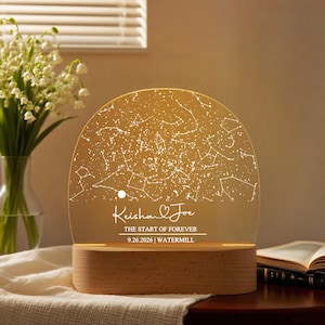 May include: A personalized night light featuring a constellation map design. The light has a wooden base and a clear, arched top with a constellation pattern and the text "Keisha & Joe, The Start of Forever, 9.26.2026, Watermill."