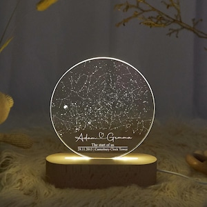 May include: A round, clear acrylic night light with a star map design. The base is made of wood and emits a warm glow. The text on the light reads "Adam & Gemma", "The start of us", and the date and location.