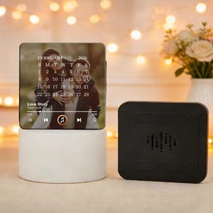 May include: A personalised photo calendar featuring a couple, the month of February 2026, and the song "Love Story". The calendar is on a white cylindrical base, with a black speaker beside it.