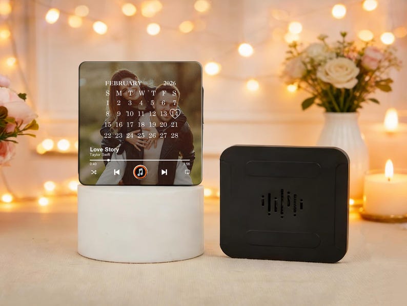 May include: A personalised photo calendar featuring a couple, the month of February 2026, and the song "Love Story". The calendar is on a white cylindrical base, with a black speaker beside it.