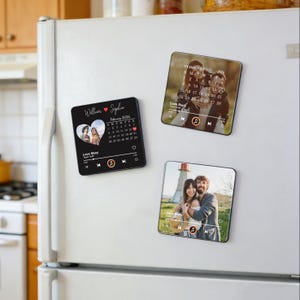 May include: Three square magnets with personalized photos and music player interfaces are displayed on a white refrigerator. Each magnet features a couple's photo, a calendar, and the text "Love Story". The magnets are black with white and red accents.
