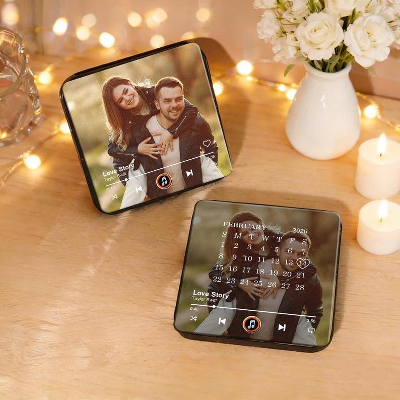 May include: Two black square devices with photos of a couple, resembling music players. One displays a photo with the text "Love Story" and a music player interface. The other shows a calendar for February 2026. The devices are on a wooden surface.