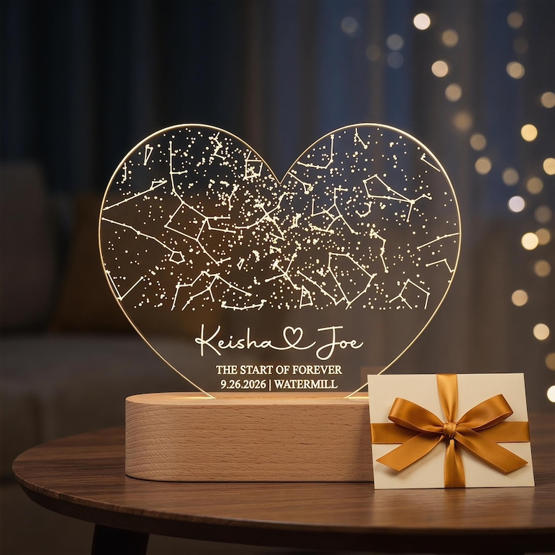May include: A heart-shaped, illuminated night light with a constellation design. The light is engraved with the names "Keisha & Joe", the date "9.26.2026", and the word "WATERMILL". It sits on a wooden base, with a gift card tied with a gold ribbon.