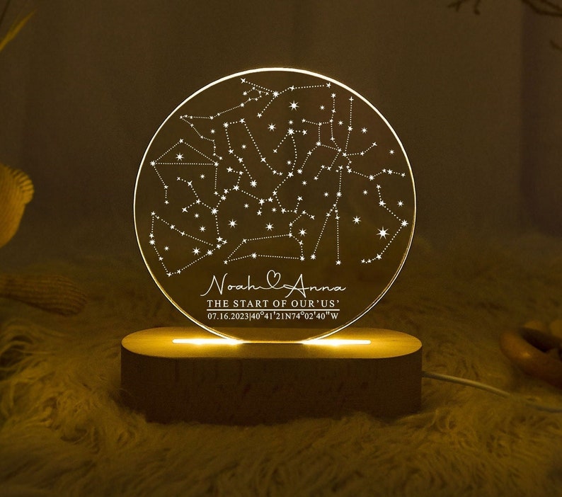 May include: A round, illuminated acrylic night light featuring a constellation map. The light sits on a wooden base and displays the text "Noah & Anna, The Start of Our Us" along with a date and coordinates. The light emits a warm glow.